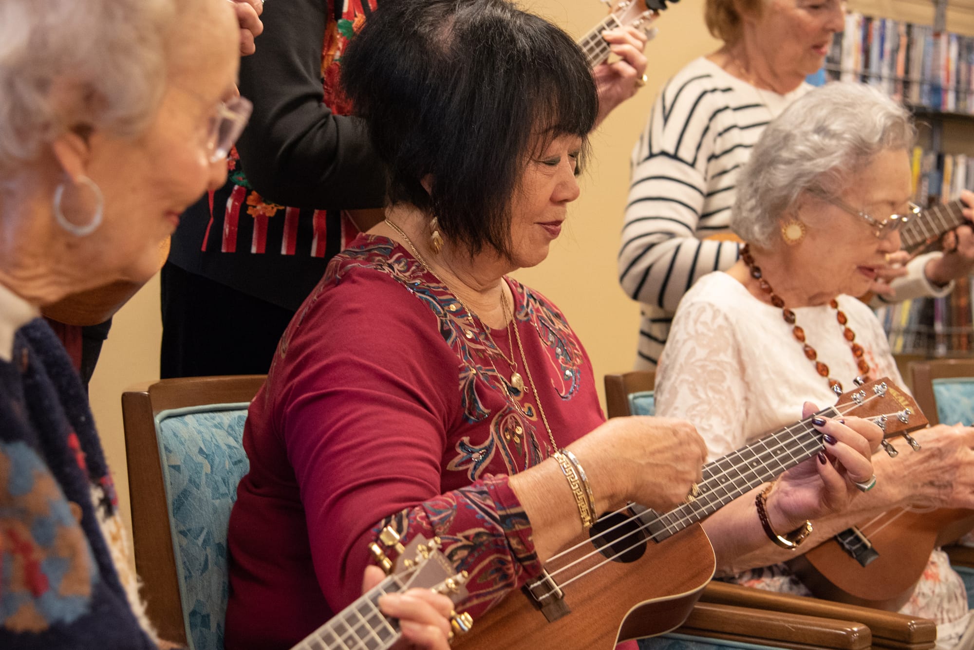 A group of elderly women sit in chairs, smiling and playing ukuleles together. Bookshelves are visible in the background, suggesting a warm, social setting.