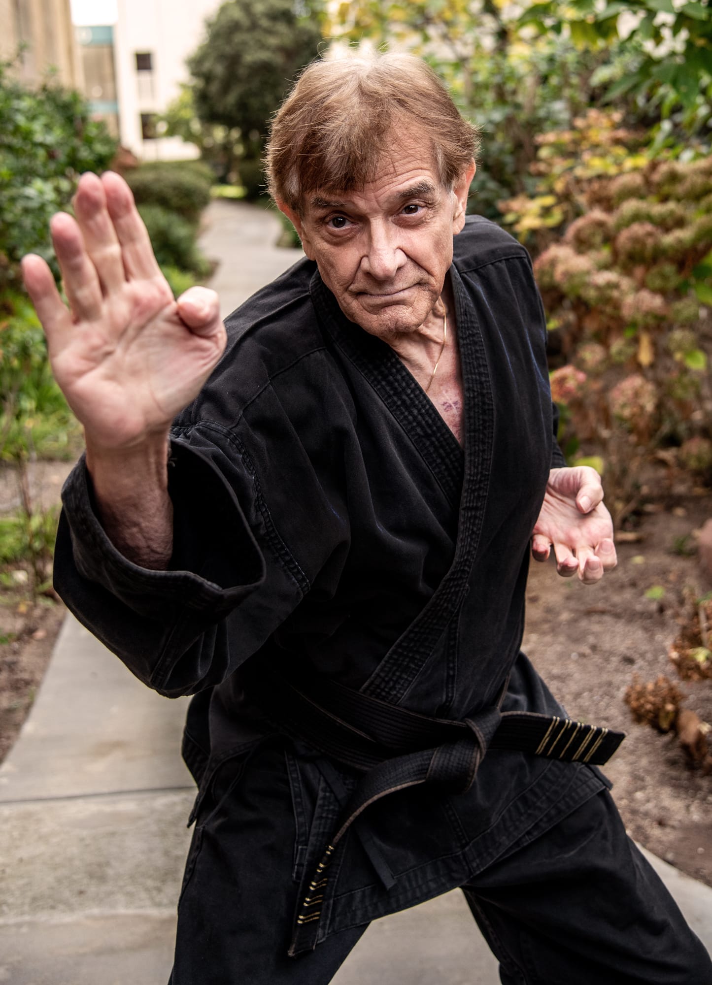 An older man wearing a black martial arts uniform and black belt strikes a defensive pose outdoors on a garden path, with one hand raised and the other near his waist.