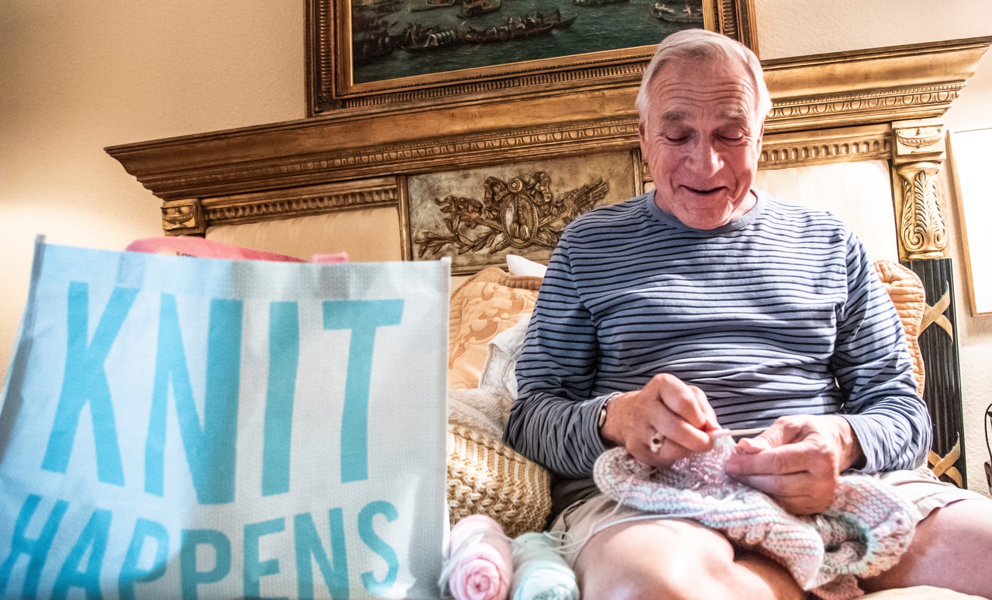 An older man sits on a couch knitting, smiling while holding yarn. Next to him is a large bag with the words KNIT HAPPENS printed in bold blue letters. Soft light fills the cozy room.