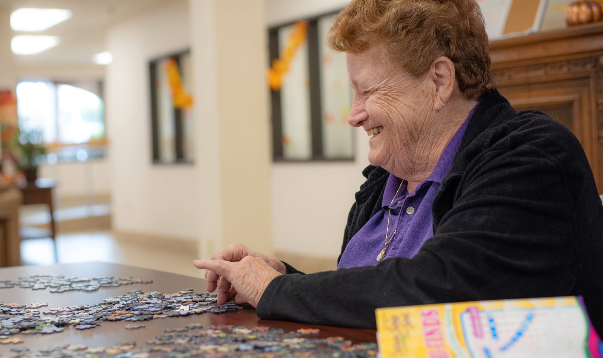 An elderly woman with short, reddish hair smiles while working on a jigsaw puzzle at a table. She wears a purple shirt and black jacket. A puzzle box is visible in the foreground.