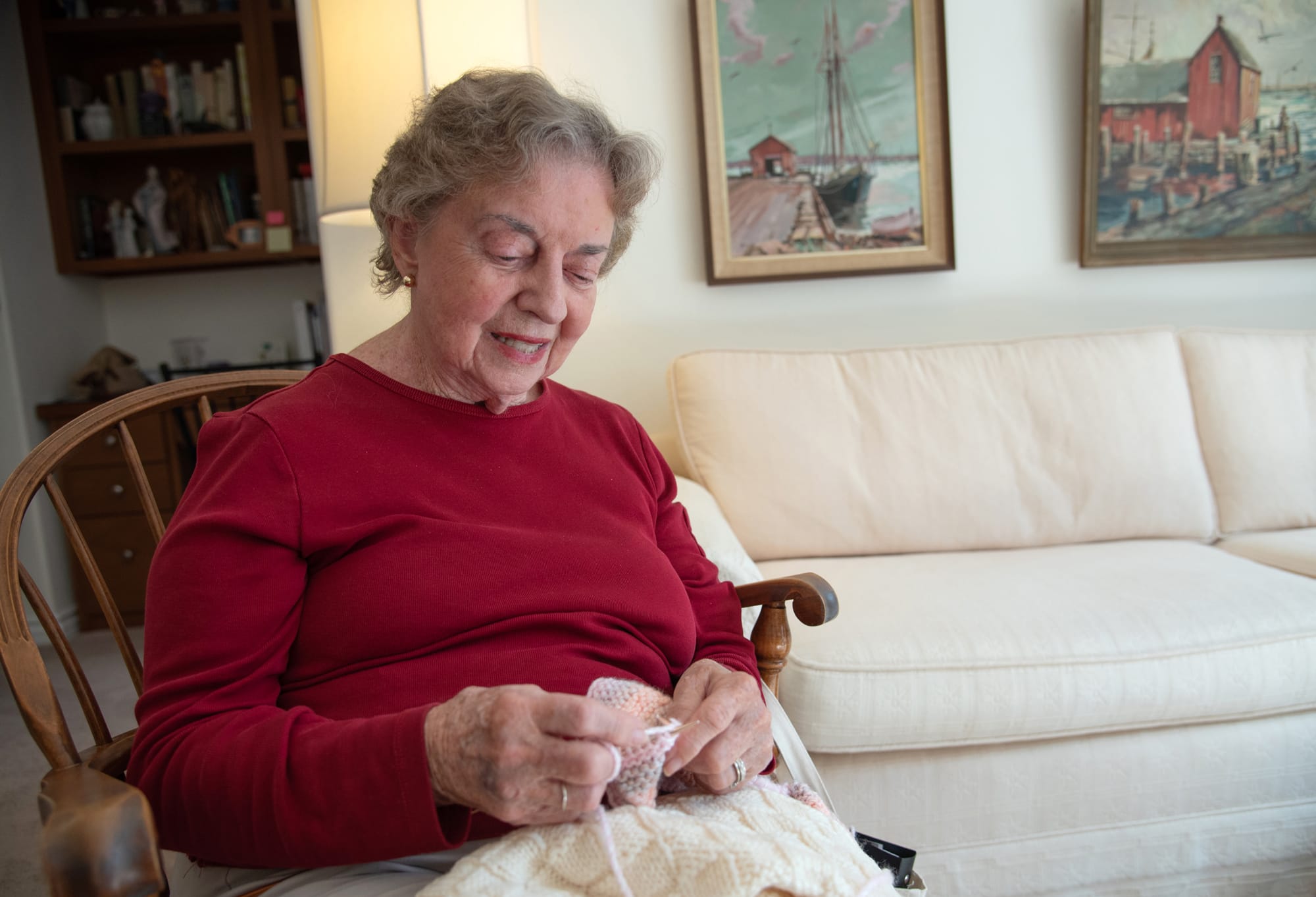 An elderly woman with short gray hair, wearing a red long-sleeve shirt, sits in a wooden chair and knits, smiling. Behind her are paintings of boats on a wall and a white couch in a cozy living room.