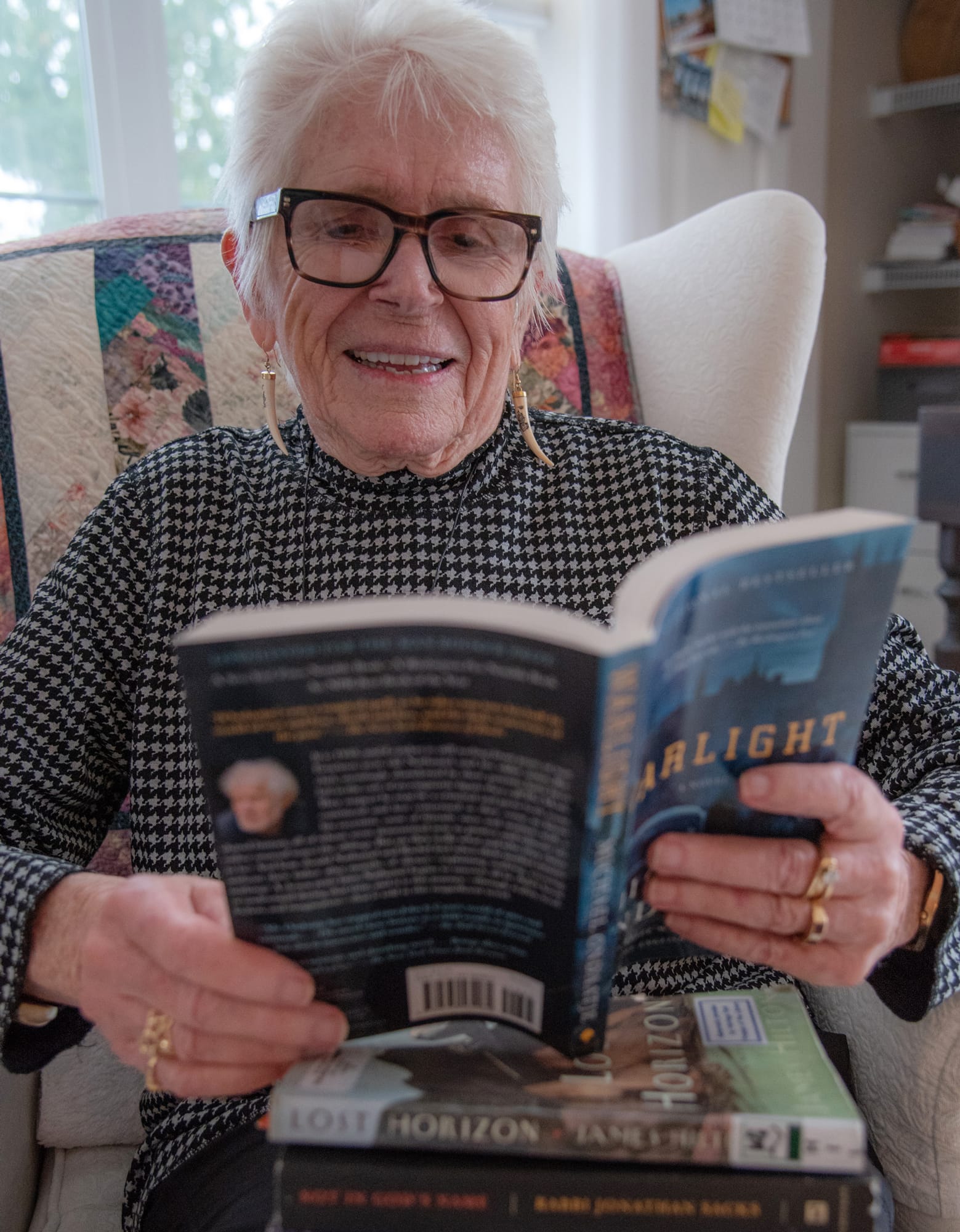 An elderly woman with short white hair and glasses sits in an armchair, smiling as she reads a book. Two more books are stacked on her lap. A colorful quilt hangs on the chair behind her.