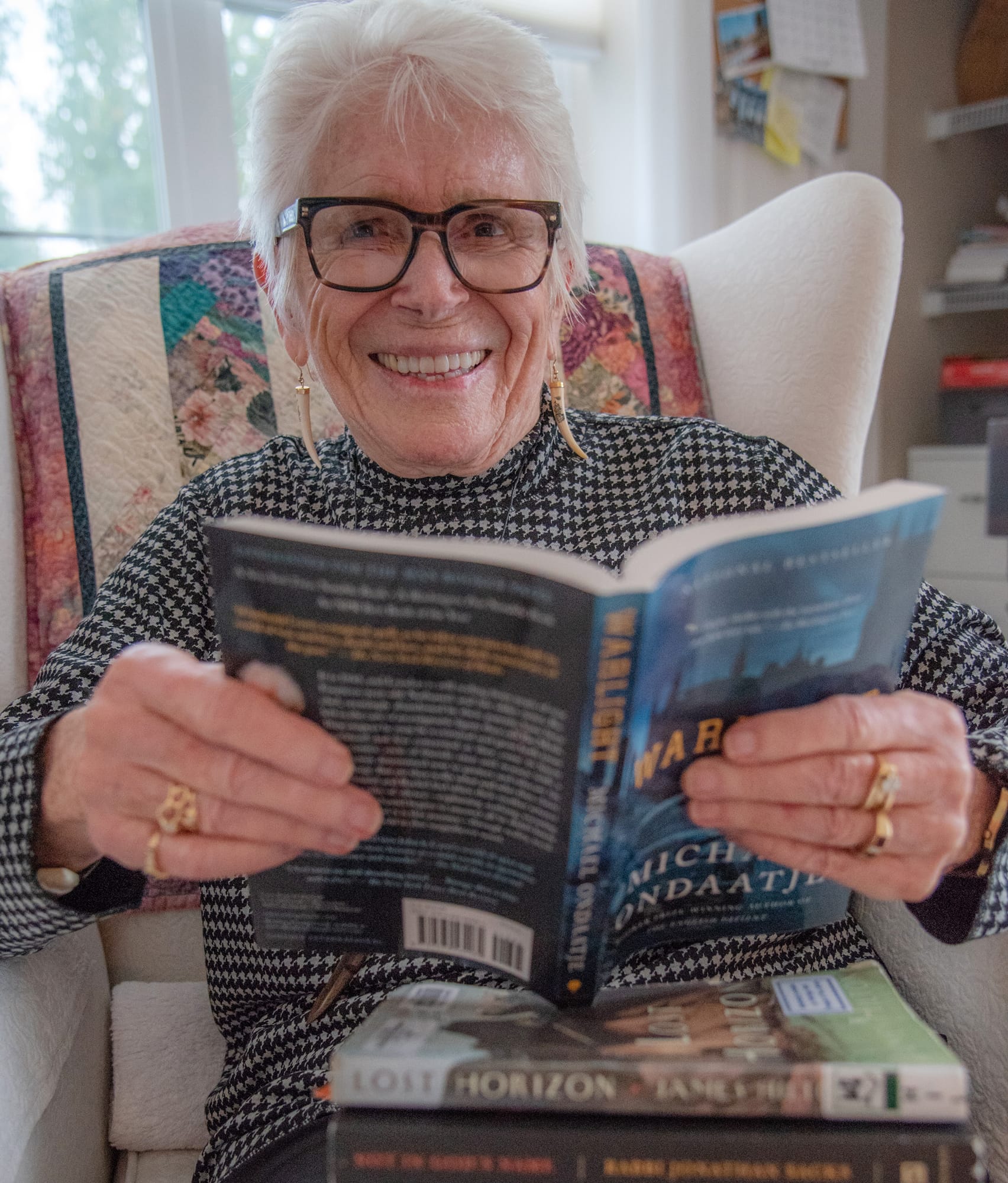 An older woman with short white hair and glasses sits in an armchair, smiling while holding an open book. Two more books are stacked on her lap. She is wearing a patterned sweater and gold jewelry.