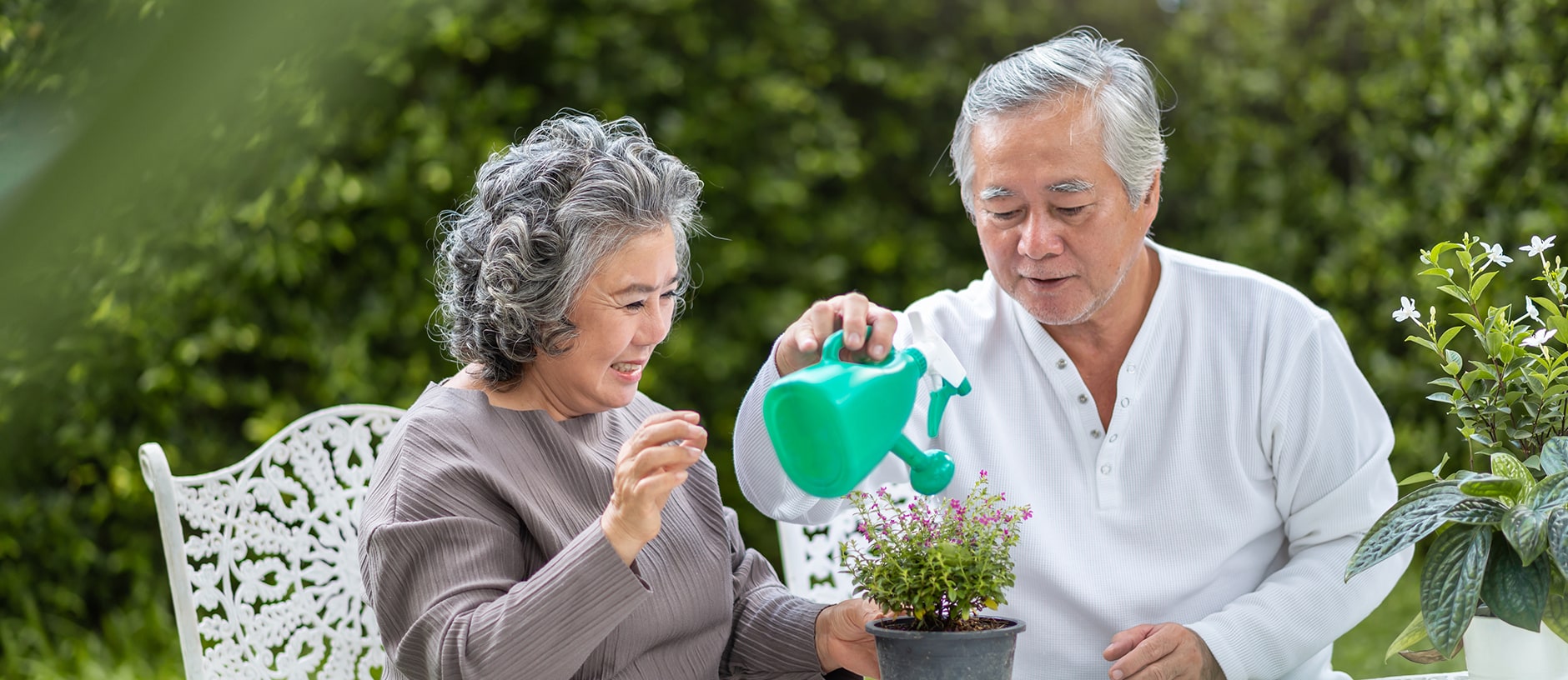 An older couple sits outdoors at a table, tending to a potted plant together. The man is watering the plant with a green watering can while the woman smiles and adjusts the leaves. Greenery is visible in the background.