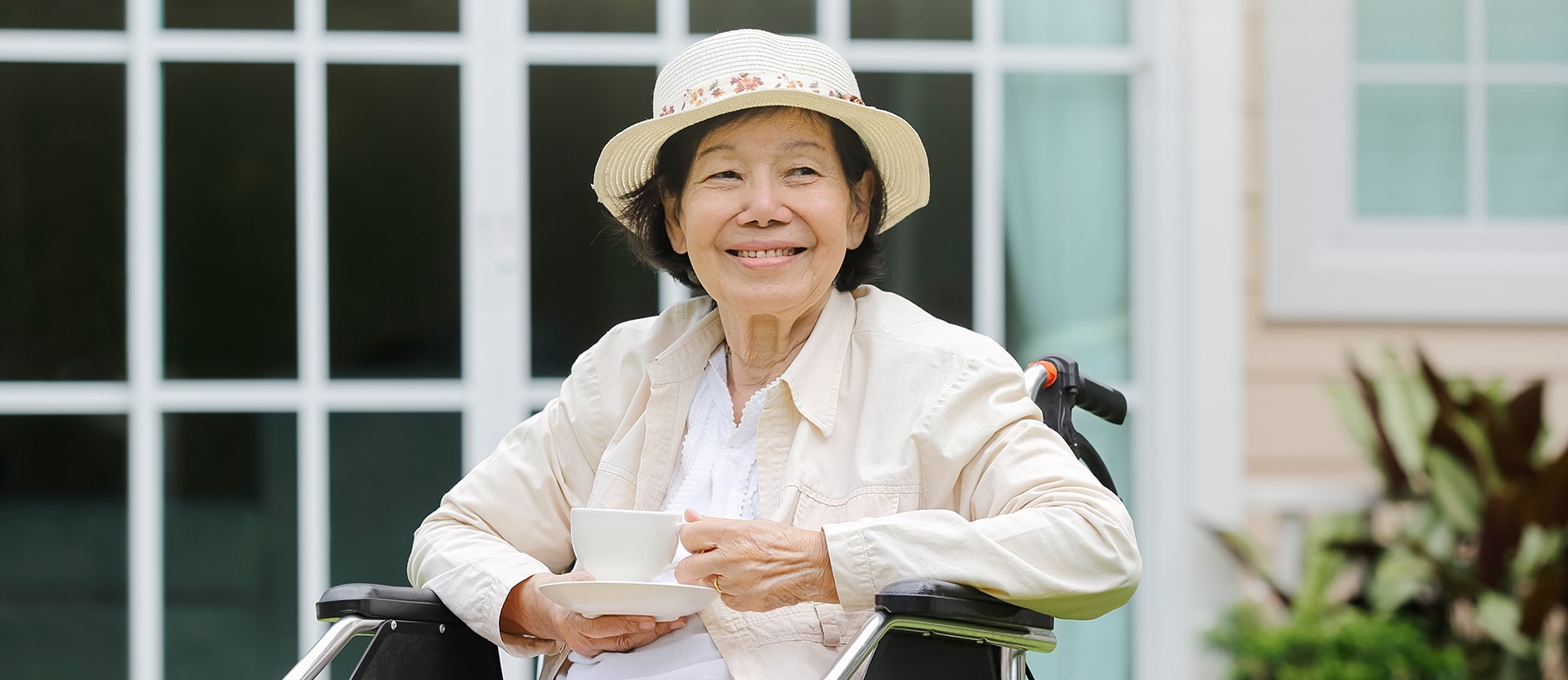 An elderly woman wearing a sun hat sits in a wheelchair outdoors, smiling and holding a cup and saucer in her hands. She is in front of a house with large windows and is dressed in light-colored clothing.