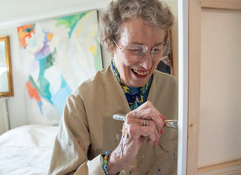 An elderly woman wearing glasses and a paint-splattered smock smiles while painting on a canvas. A colorful abstract painting hangs on the wall behind her.