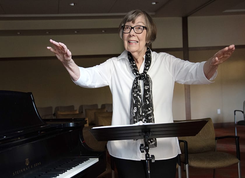 An older woman wearing glasses and a black-and-white scarf stands beside a piano, smiling and raising her arms as if conducting music, in a room with chairs in the background.