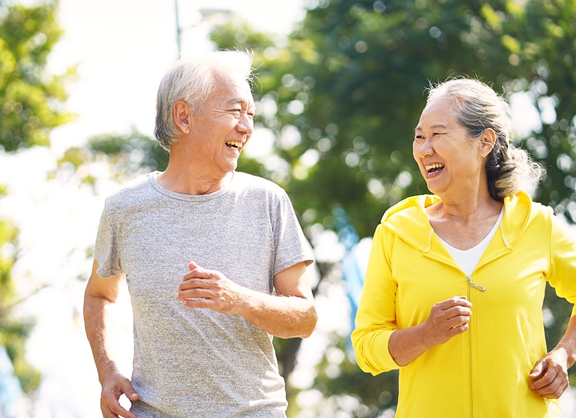 An older man and woman are jogging outdoors on a sunny day, smiling and looking at each other. They are surrounded by green trees, and both appear happy and healthy.