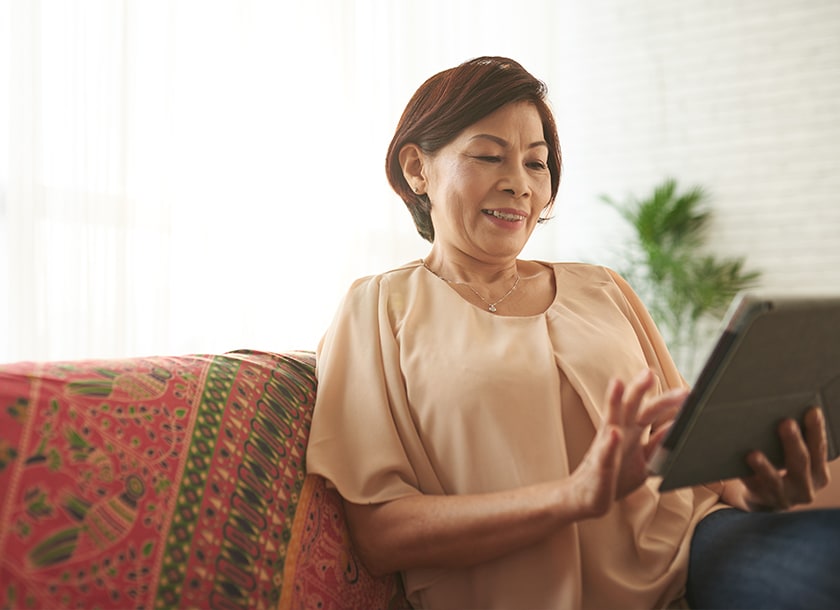 A smiling woman sits on a colorful patterned couch, using a tablet. She is relaxed and casually dressed, with natural light coming in from a window behind her and a plant in the background.