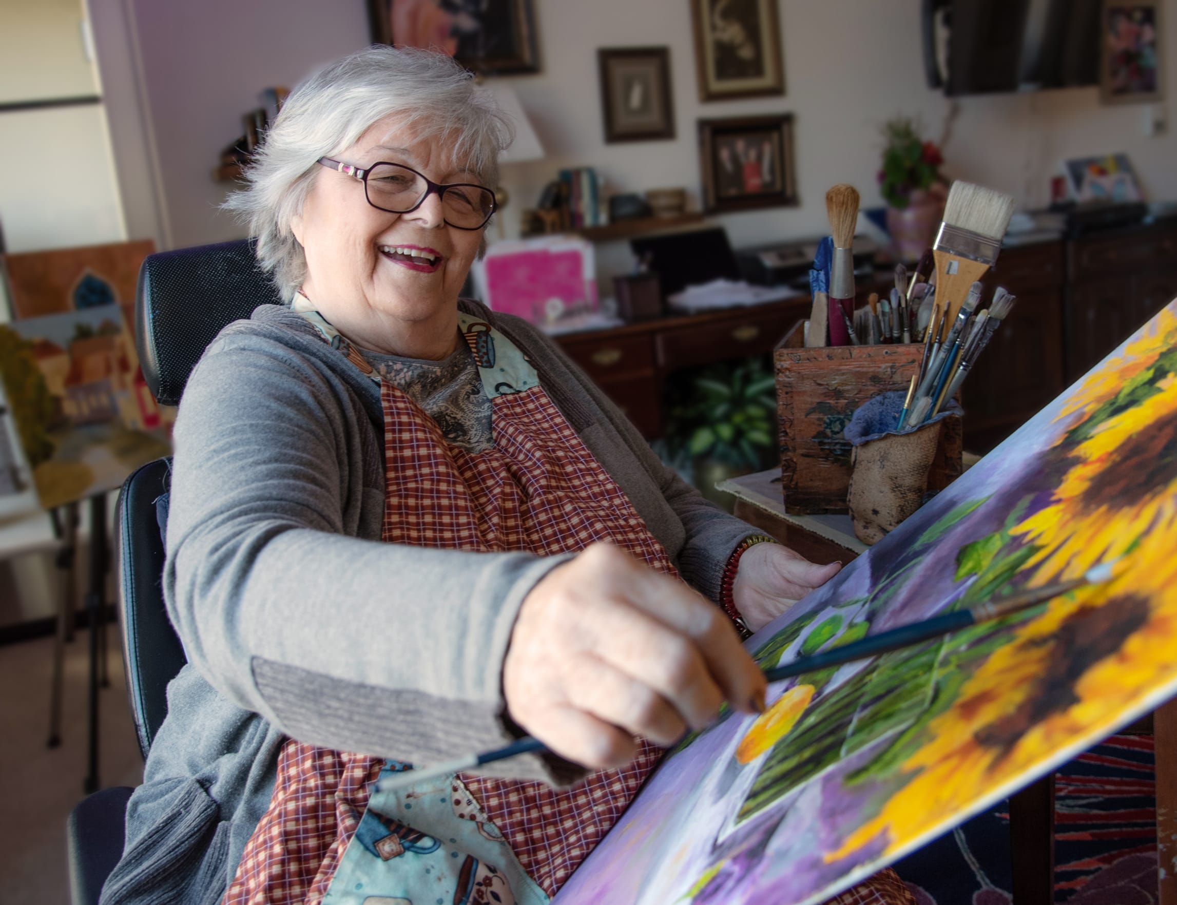 An older woman with gray hair and glasses smiles while painting a colorful sunflower artwork in a cozy, art-filled room. She wears an apron and holds a paintbrush, surrounded by art supplies and paintings.