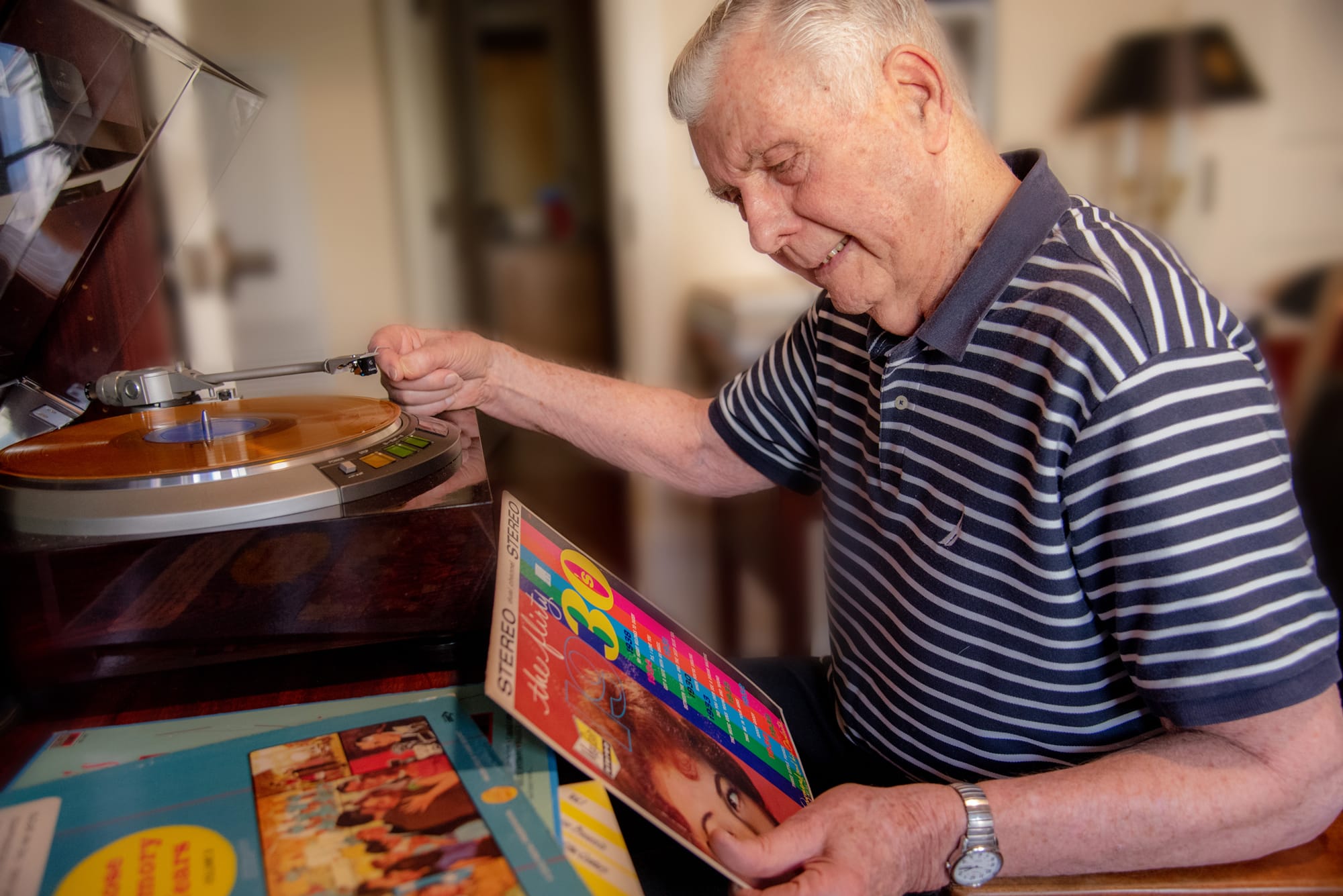 An elderly man in a striped shirt smiles while selecting a vinyl record to play on a turntable. Several colorful record covers are spread out in front of him on the table.