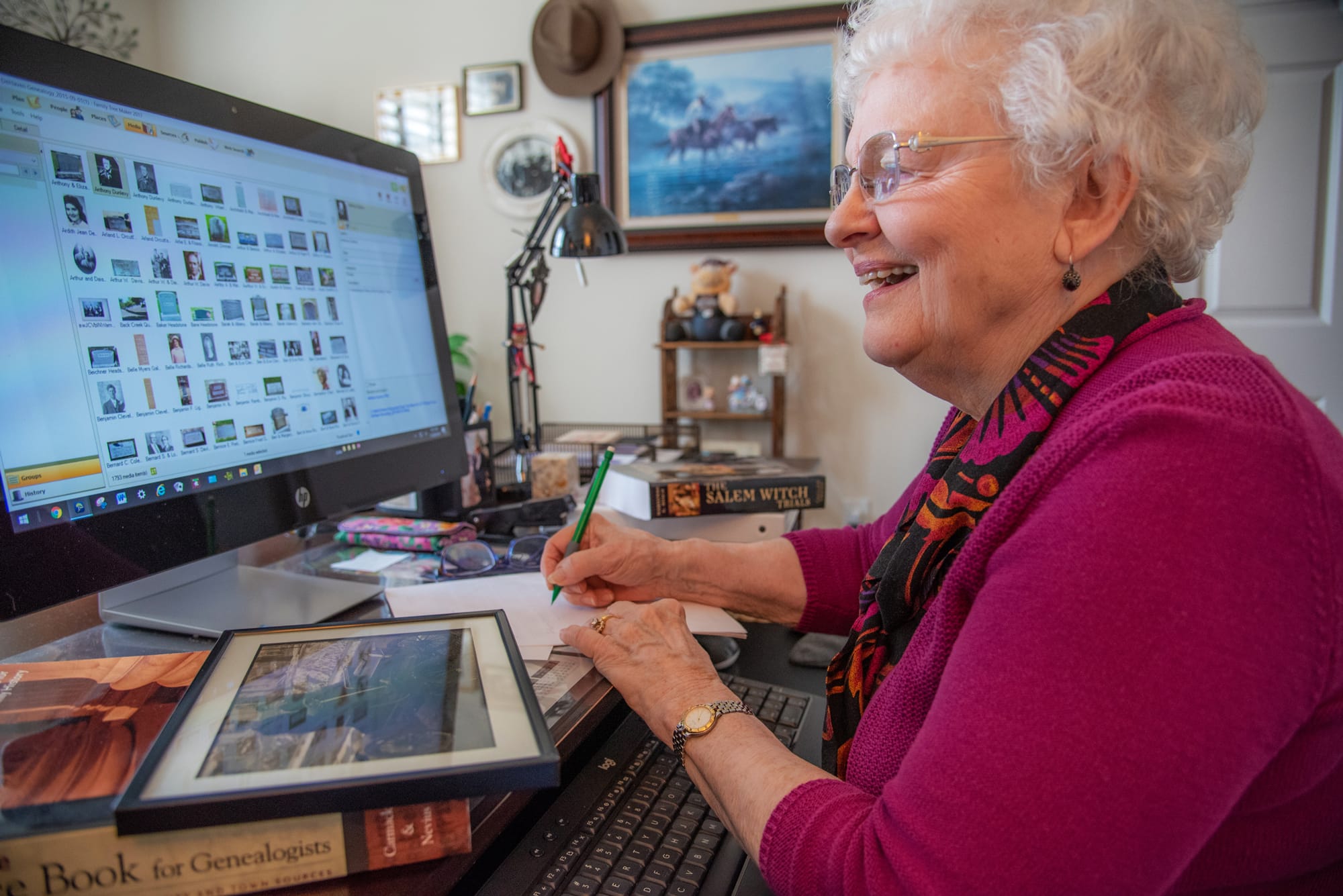 An elderly woman smiles while sitting at a desk, writing on paper beside a computer displaying photo thumbnails. Books and a framed photograph are on the desk, and office items decorate the background.