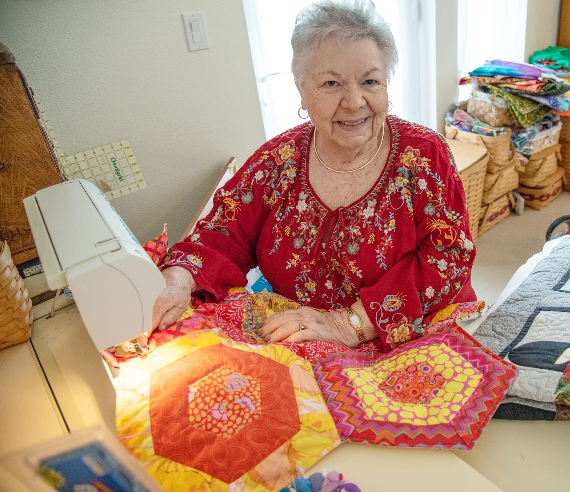 An elderly woman smiles while sewing a colorful quilt at a sewing machine, wearing a red embroidered blouse. Finished quilts and fabric are neatly stacked around her in a well-lit room.