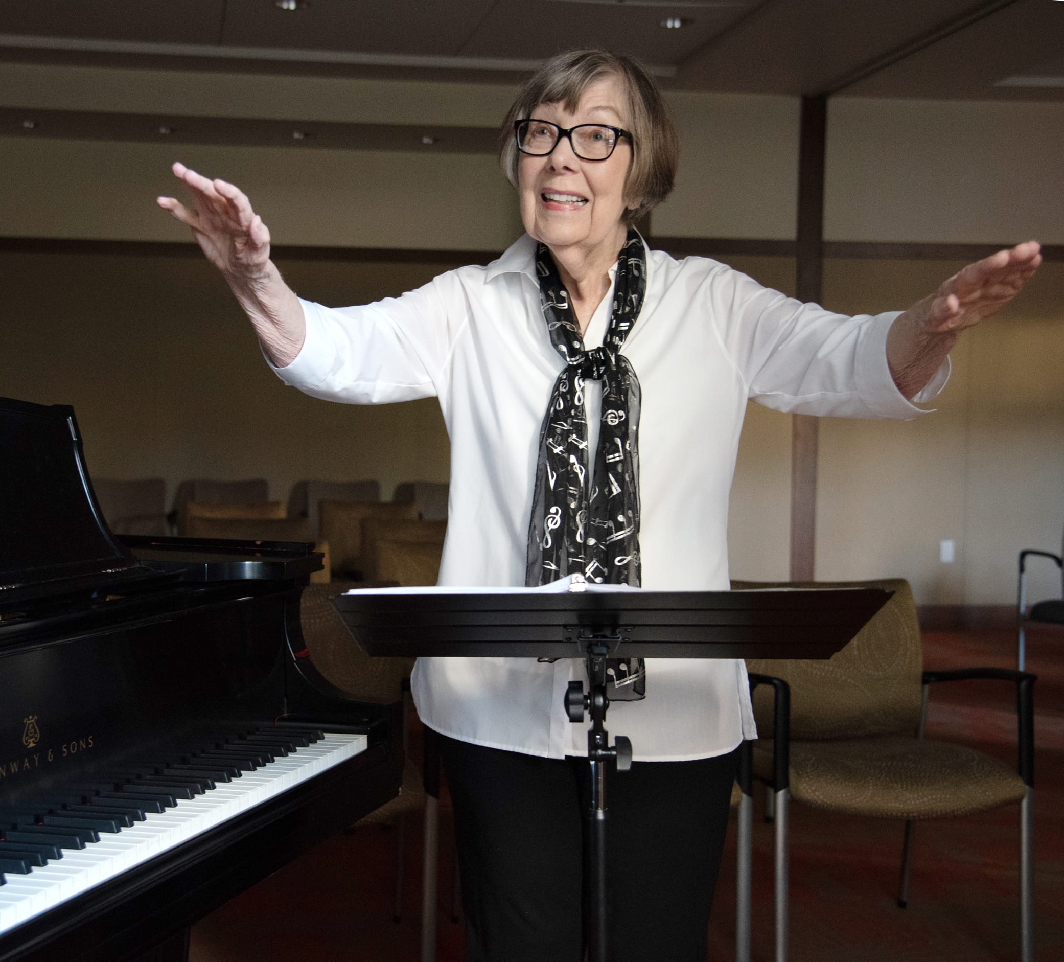 An older woman wearing glasses, a white blouse, and a music note scarf stands in front of a piano, smiling and raising her arms as if conducting in a room with empty chairs.