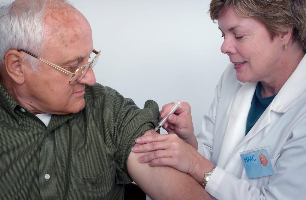 An older man receives a covid 19 vaccination in his upper arm from a healthcare worker wearing a white coat and name badge. The man watches as the healthcare worker carefully administers the shot.