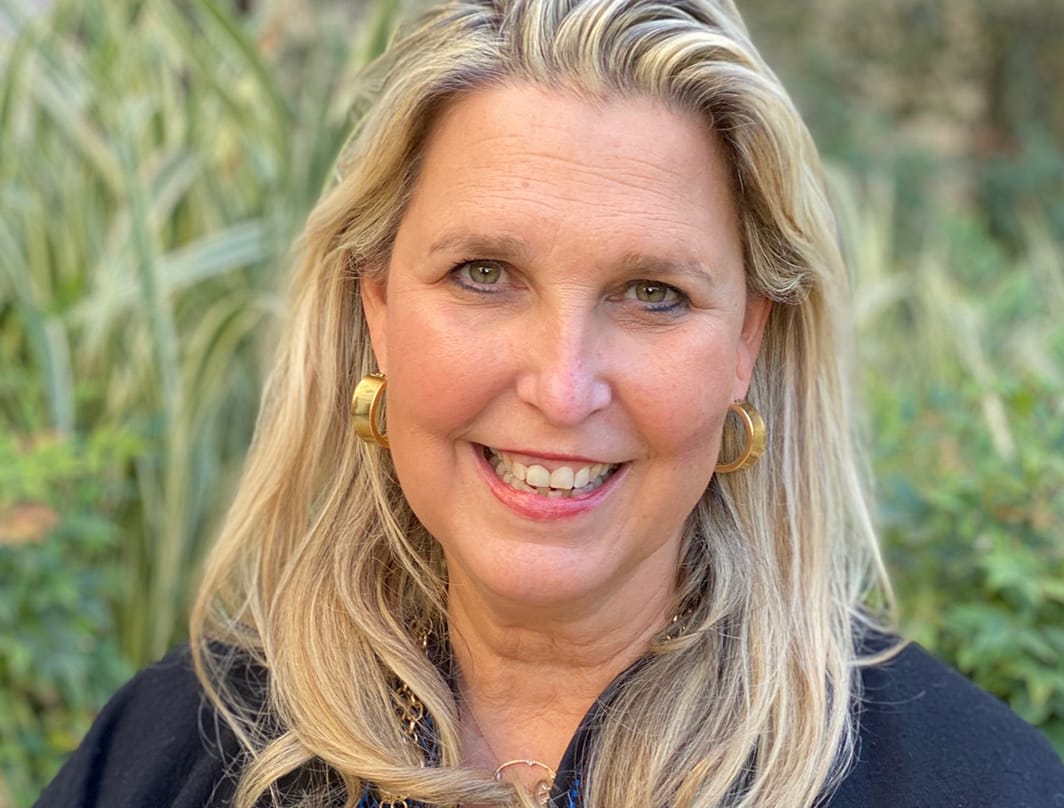 Elizabeth Saldebar, a middle-aged woman with long blonde hair, smiles at the camera. She is wearing gold hoop earrings and a dark top, standing outdoors with green plants in the background.