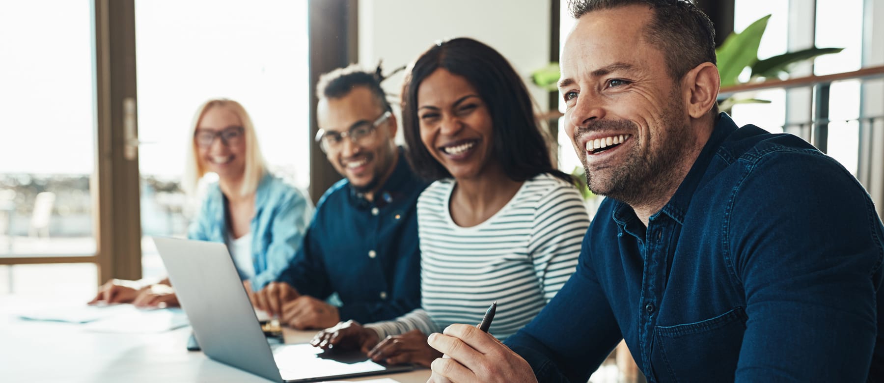A group of four smiling adults sit at a table with a laptop in a bright office, appearing engaged and collaborative.