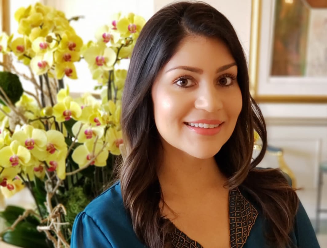Lizett Leandro, with long brown hair and a blue blouse, smiles in front of a large bouquet of yellow orchids in a bright, elegant room.