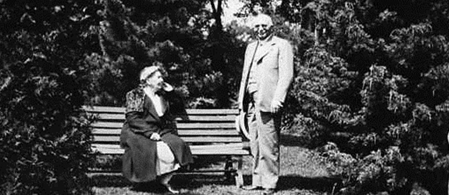 An older woman sits on a wooden bench outdoors, wearing a dress and coat, while an older man in a suit stands next to her. They are surrounded by trees and greenery, with sunlight casting shadows.