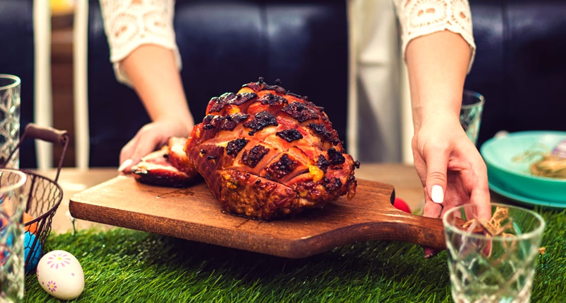 A person sets a wooden board with a glazed, baked ham garnished with fruit on a table decorated with artificial grass and pastel-colored Easter eggs. Glasses and plates are nearby.