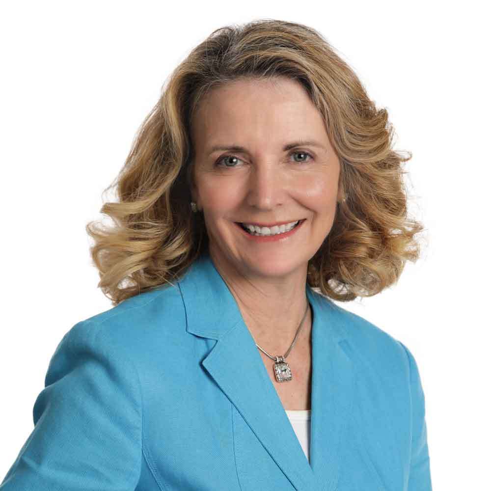 Smiling middle-aged woman with wavy blonde hair, wearing a light blue blazer over a white top and a silver necklace, posing against a plain white background.