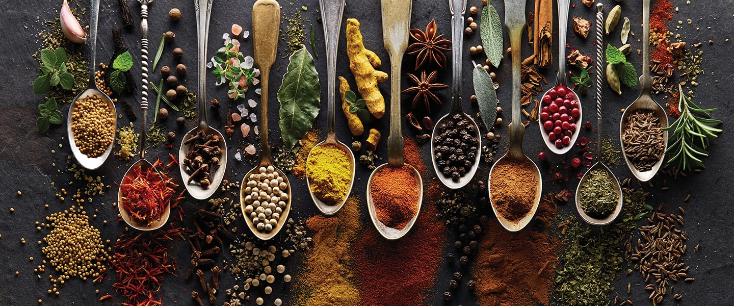 ECS_Events_Delight-the-Senses_spices A variety of metal spoons filled with colorful spices and herbs, including turmeric, peppercorns, star anise, and bay leaves, arranged in a row on a dark surface with scattered loose spices around them.