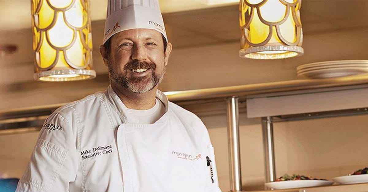 A smiling chef in a white uniform and hat stands in a restaurant kitchen with hanging pendant lights and prepared dishes visible on the counter behind him.