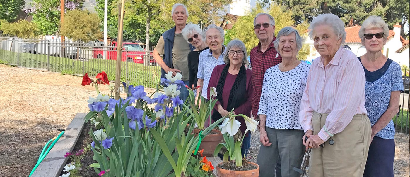 A group of older adults stands and smiles together beside a garden bed filled with colorful blooming flowers on a sunny day. Trees and a fence are visible in the background.