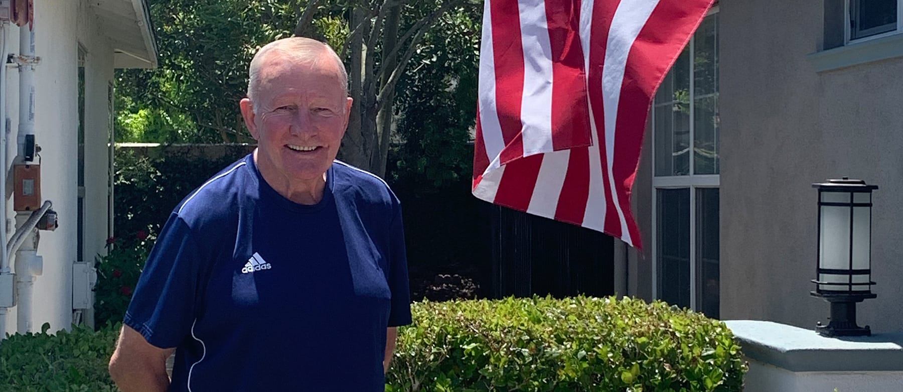 An older man in a navy Adidas shirt stands outside next to green bushes. An American flag hangs nearby, and a house with a window and outdoor lamp is in the background. The weather is sunny.
