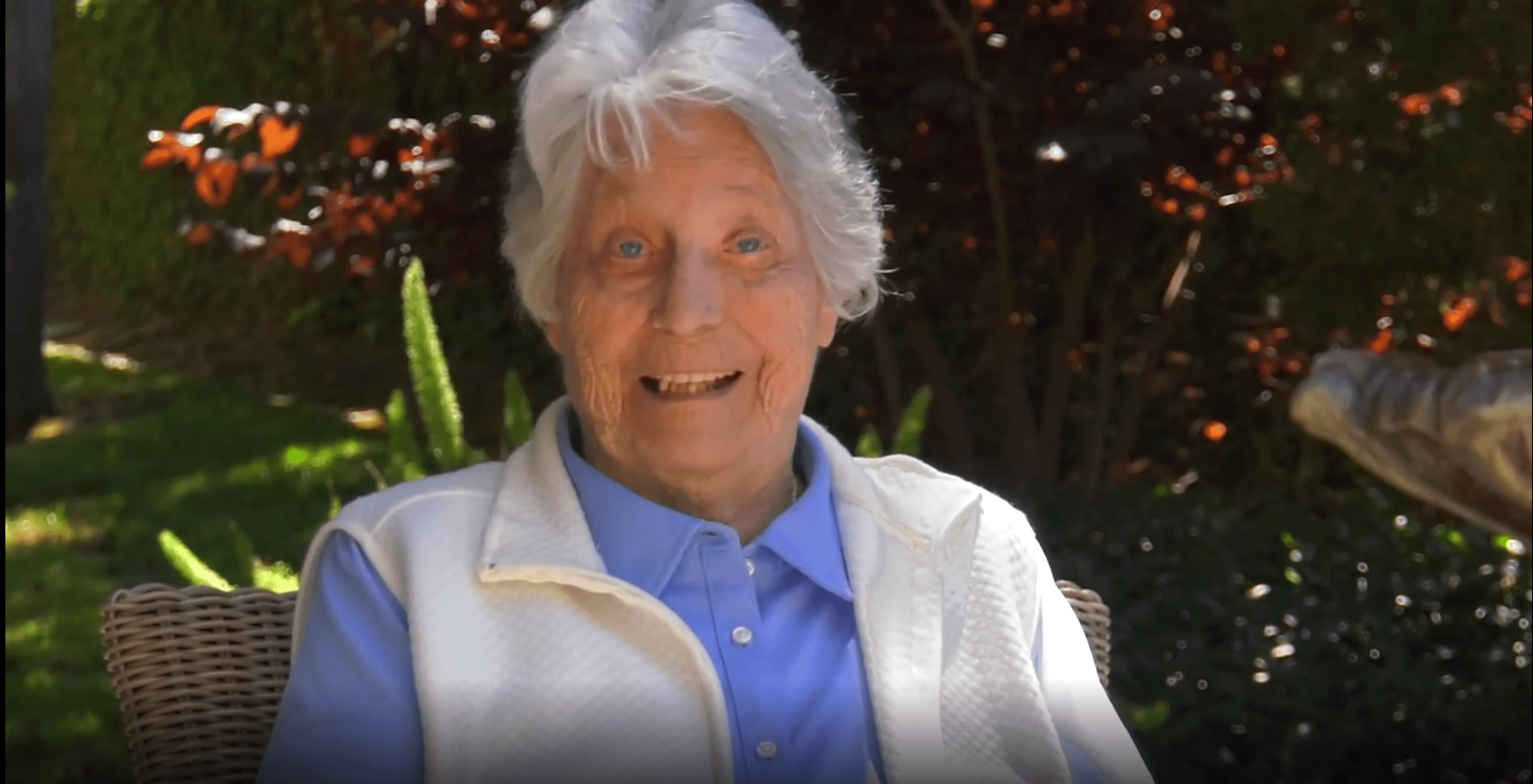 An elderly person with white hair, wearing a light blue shirt and white vest, sits outdoors on a sunny day, smiling at the camera with greenery and red-leaved plants in the background.