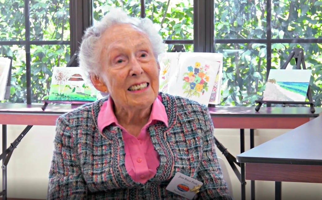 An elderly woman with curly gray hair, wearing a plaid jacket over a pink blouse, sits smiling in front of a table displaying colorful paintings, with large windows and greenery in the background.