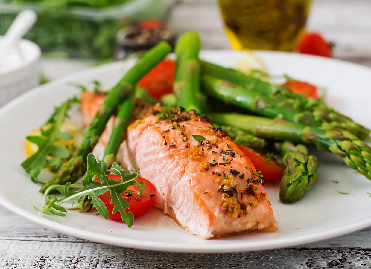 A plate of cooked salmon garnished with herbs, served with asparagus, cherry tomatoes, arugula, and lemon wedges on a white plate. A blurred salad and olive oil are in the background.