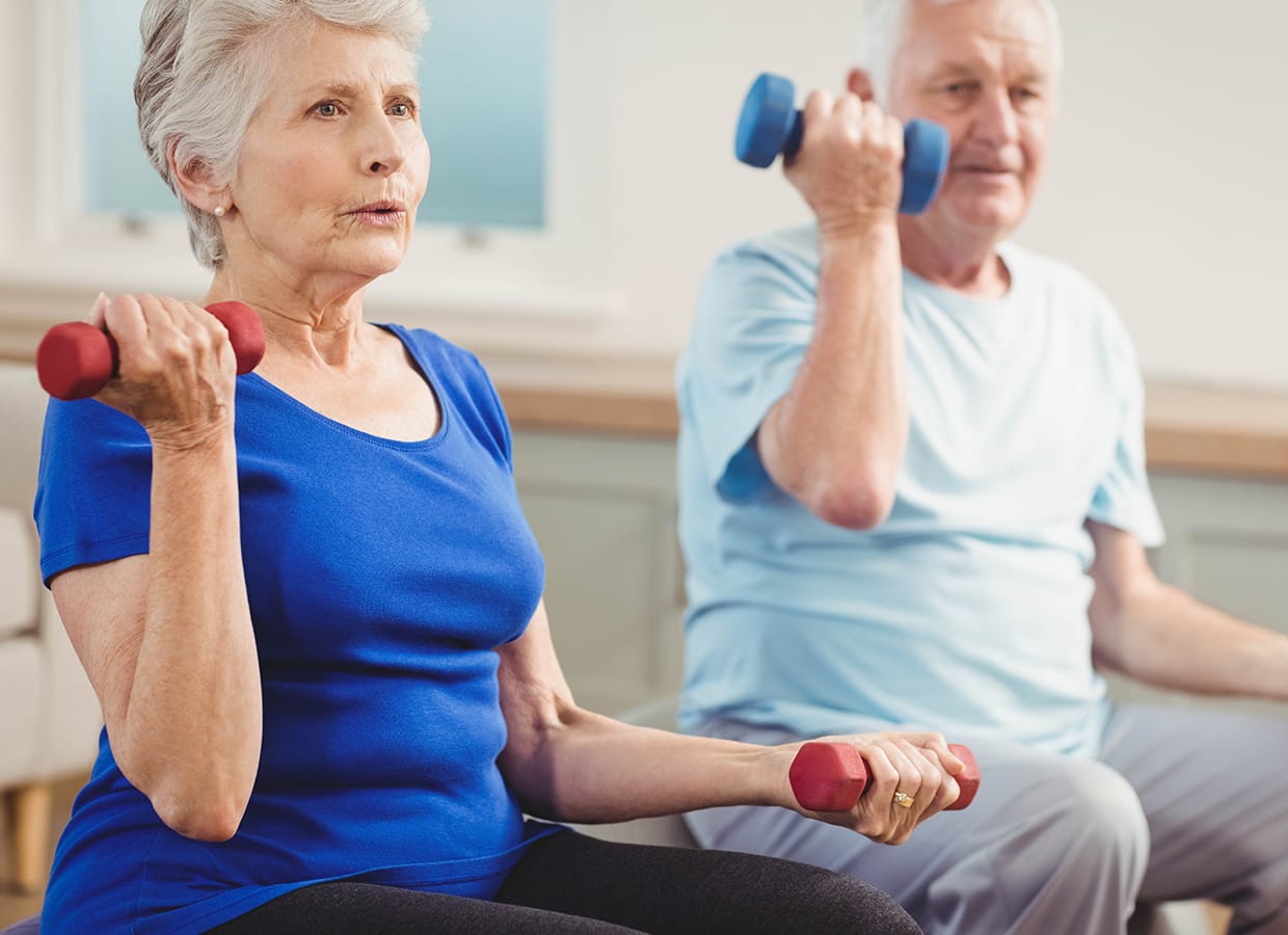Two older adults sitting indoors, exercising with dumbbells. The woman in the foreground wears a blue shirt, and the man behind her wears a light blue shirt. Both are lifting weights with focused expressions.
