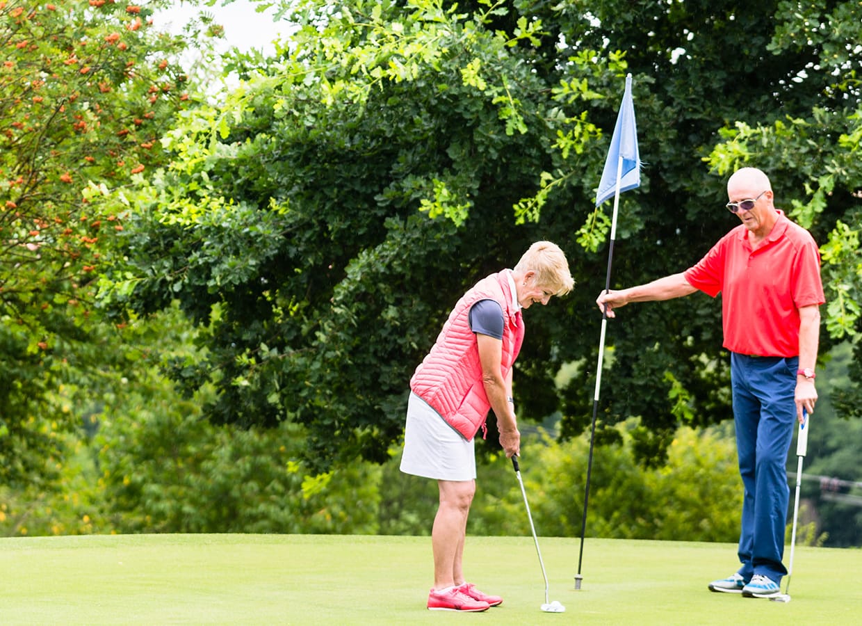 An older woman prepares to putt on a golf green while a man in sunglasses and red shirt holds the flagstick nearby. Trees and greenery fill the background on a sunny day.