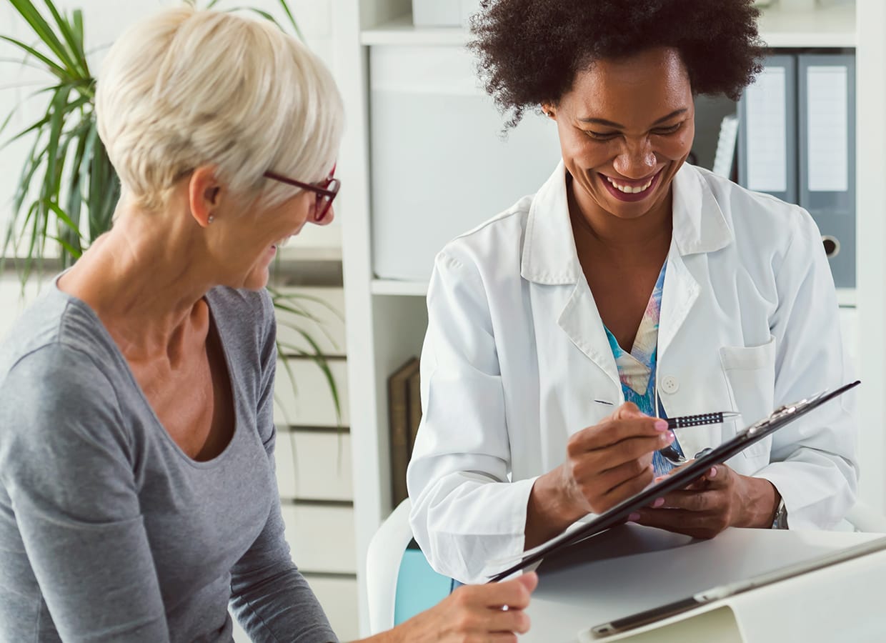 A doctor smiles while showing a clipboard to a patient with short gray hair and glasses. Both women appear engaged and happy in a brightly lit medical office.