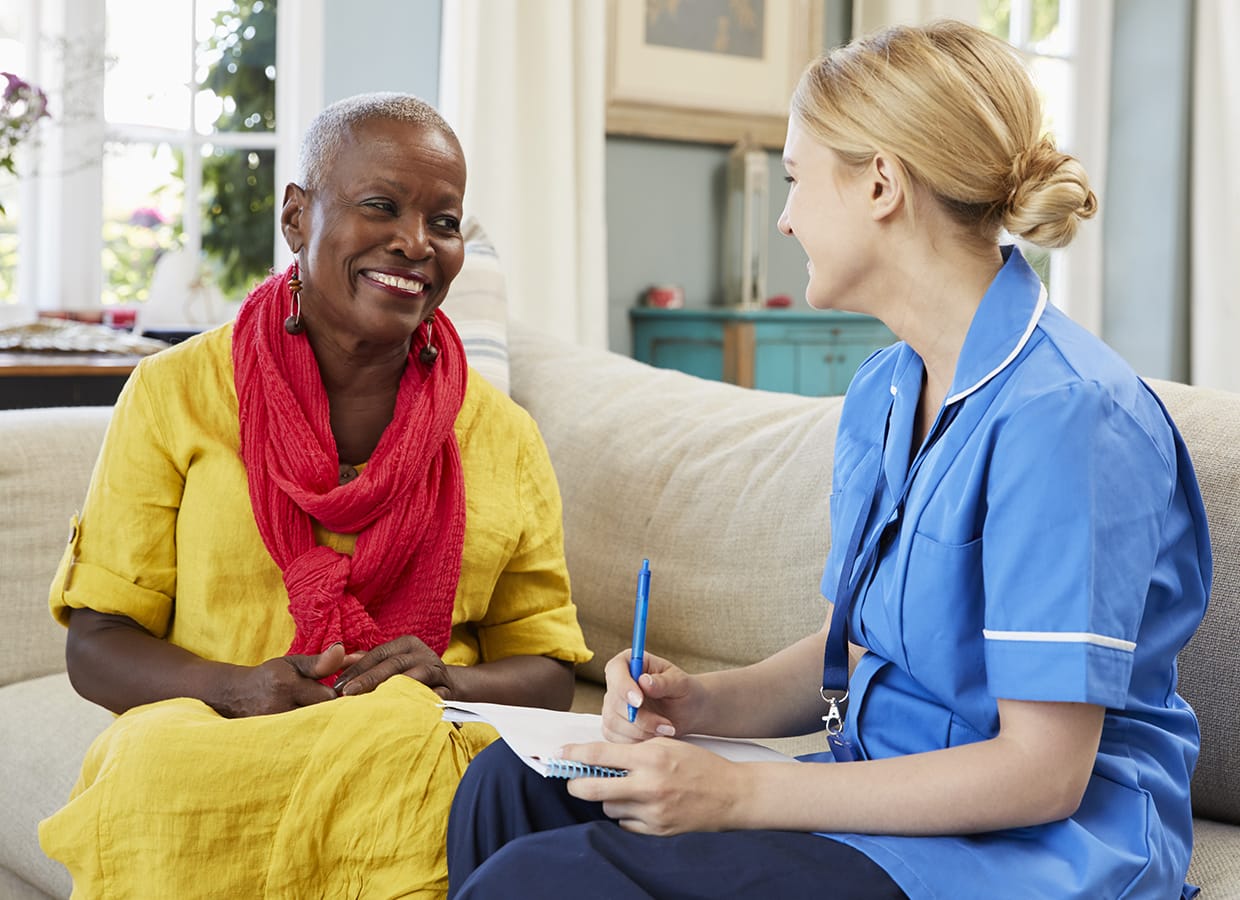 An older woman in a yellow dress and red scarf smiles while talking with a nurse in blue scrubs, who is writing on a clipboard. They are sitting on a couch in a bright, homey living room.