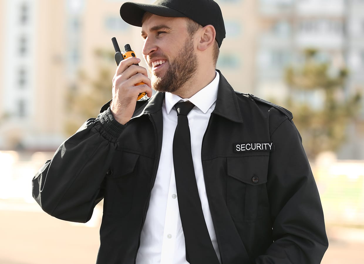 A smiling security guard in a black uniform and cap speaks into a walkie-talkie outdoors, with blurred buildings and trees in the background.