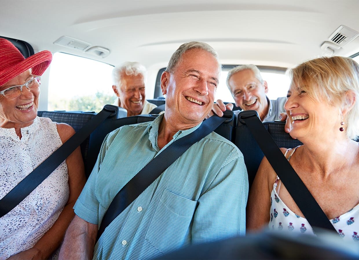 Five older adults sit in a car, smiling and laughing together, their creativity shining as they share stories. Three are seated in the back with seatbelts, while two are in front—all enjoying each other's company.