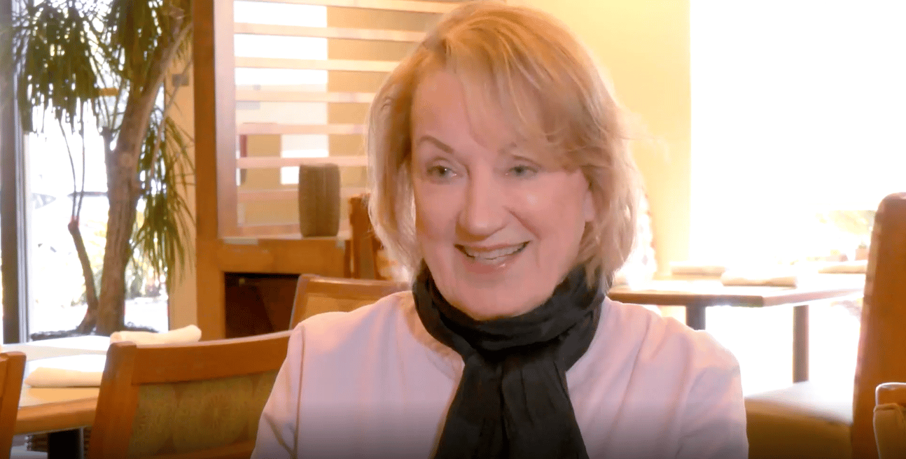 A woman with blonde hair wearing a light pink top and dark scarf smiles while sitting in a warmly lit restaurant with wooden furniture and soft natural light.