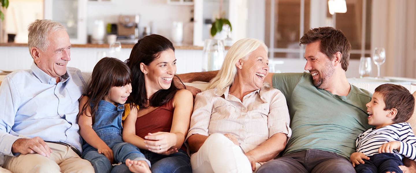 Three generations of a family—two older adults, two parents, and two young children—sit together on a couch, smiling and laughing in a bright, cozy living room.