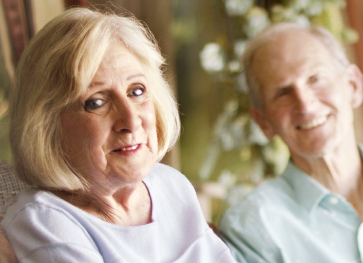 An elderly woman with light blonde hair smiles gently at the camera, while an elderly man with short gray hair sits beside her, also smiling. The background is softly blurred with hints of greenery.