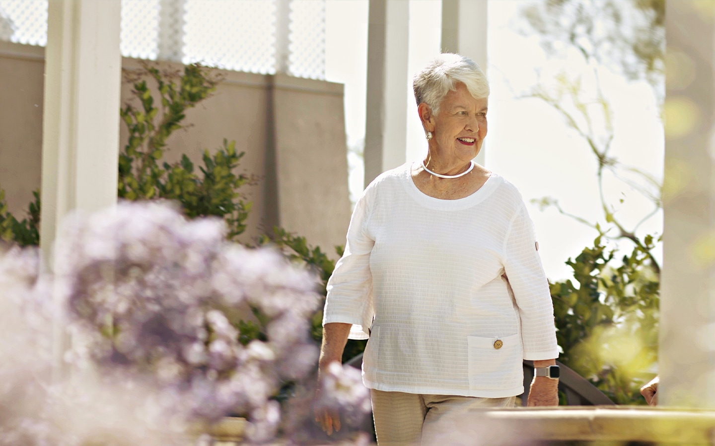 An older woman with short gray hair, wearing a white long-sleeve top and beige pants, walks outdoors near greenery and flowers, smiling and enjoying the sunny day.