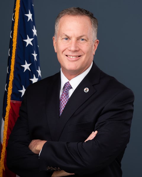 A man in a dark suit and patterned tie stands smiling with arms crossed in front of a U.S. flag, against a plain blue-gray background.