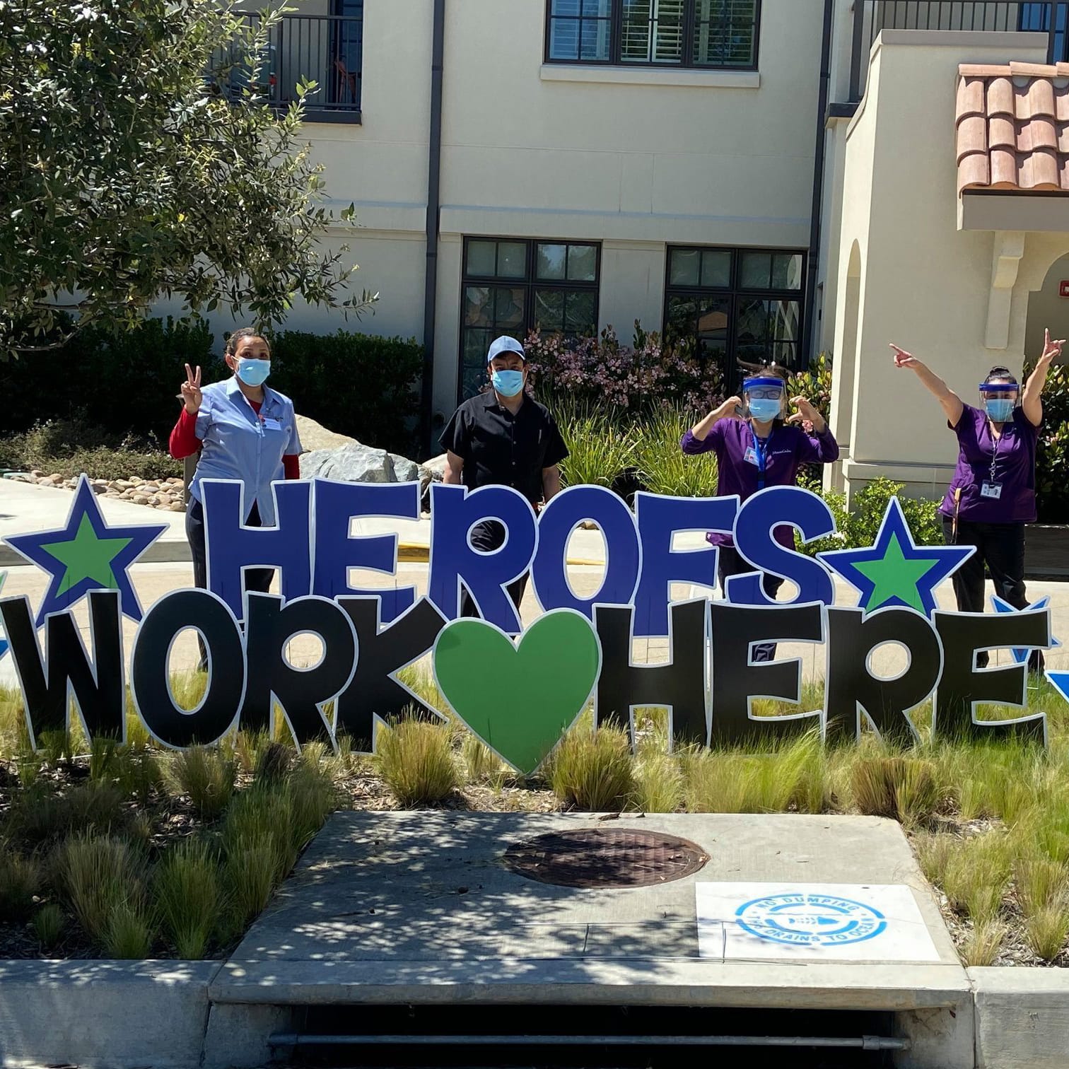 Three people in scrubs and masks stand outside a building, smiling and making peace signs behind a large sign that reads HEROES WORK HERE with stars and a green heart.