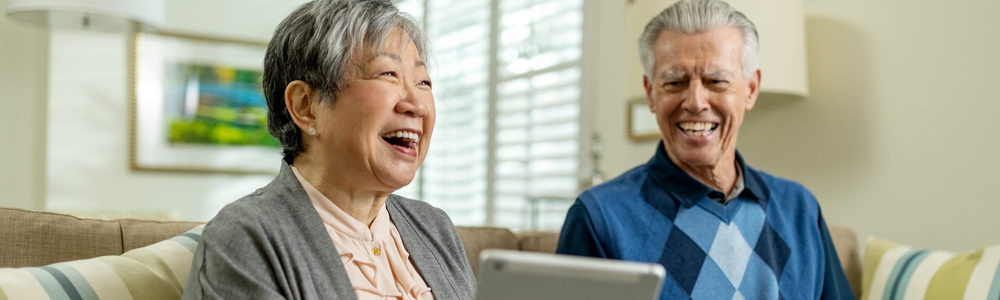 An elderly woman and man sit together on a couch, smiling and laughing while looking at a tablet in a bright, cozy living room.