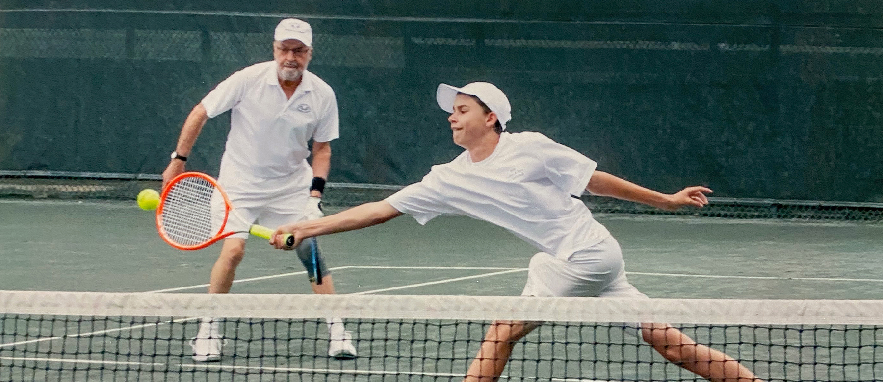 Two men in white tennis outfits play doubles on an outdoor court. One stretches forward to hit the ball with his racket, while the other stands behind him, ready and watching the action.
