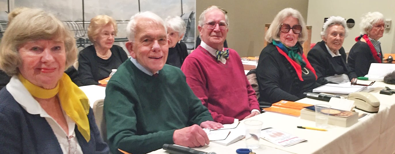 A group of older adults sit side by side at a conference table, smiling at the camera. The table is covered with papers, books, and glasses. Most are dressed warmly with scarves and sweaters.