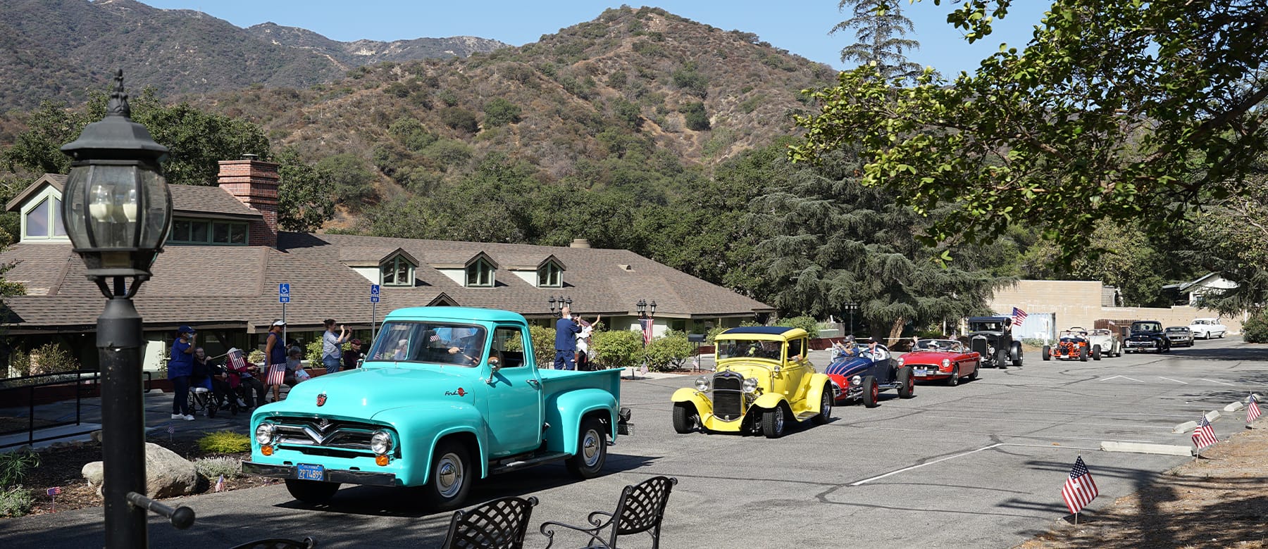 Several vintage cars, including a bright blue pickup and a yellow coupe, are parked along a tree-lined street with mountains in the background and people admiring the vehicles.