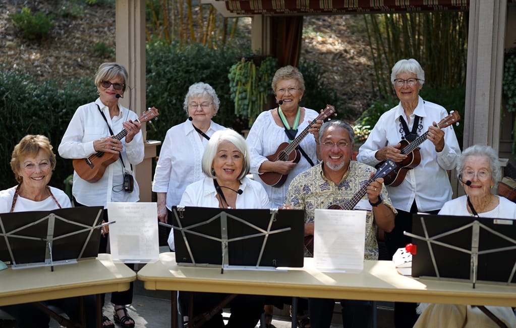 A group of smiling older adults, mostly women, play ukuleles and sing together outdoors. Some are seated behind music stands, while others stand behind them. They wear white shirts and appear to be enjoying the performance.