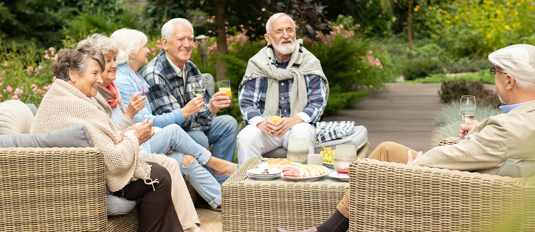 A group of six older adults sit on wicker chairs outdoors, smiling and chatting while holding drinks. There are snacks on a table in front of them, and they are surrounded by greenery and flowers.